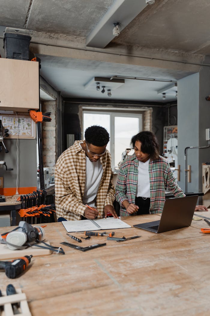 Two artisans collaborate in a woodworking workshop. Tools and laptop on the workbench.