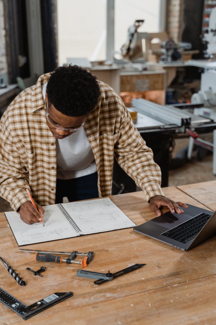 A craftsman sketches designs while working in a woodshop with tools and a laptop nearby.